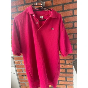 Rosa pikétröja Lacoste XXL - Snygg rosa pikétröja från Lacoste i storlek XXL. Klassisk krage med två knappar och den ikoniska krokodilloggan broderad på bröstet. Tröjan är i bomull och har korta ärmar.