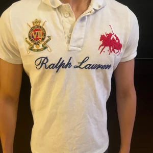 Vit piké från Ralph Lauren - Vit pikétröja från Ralph Lauren med klassisk krage, broderad röd polospelare på bröstet och stort blått Ralph Lauren-tryck. Snygg broderad patch med klubbkänsla på vänster sida. Perfekt för en sportig och stilren look. Jävligt nice pike men har tyvärr växt ur den, nypris ca 1200 