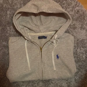 Grå hoodie från Polo Ralph Lauren - Snygg ljusgrå hoodie från Polo Ralph Lauren med dragkedja, huva med snörning och klassisk blå broderad logga på bröstet. Tröjan har känguruficka och är tillverkad i mjuk bomullsmix. Perfekt för chill dagar eller när du vill ha en clean look. Storlek M men sitter som en S/XS. Hör av dig vid frågor eller för fler bilder! 