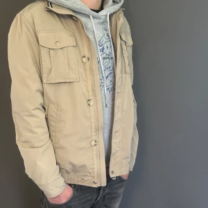 Beige jacka från J.Lindeberg, storlek L - Snygg beige jacka från J.Lindeberg med klassisk look. Jackan har flera fickor framtill, både med knapp och dragkedja, samt stängs med både dragkedja och knappar. Tillverkad i polyester och har meshfoder i grönt och prickigt tyg på insidan. Perfekt för vår och höst.