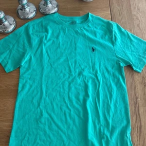 Turkos t-shirt från Polo Ralph Lauren - Snygg turkos t-shirt från Polo Ralph Lauren med klassisk passform och korta ärmar. T-shirten har en liten broderad logga på bröstet och är tillverkad i mjuk bomull. Perfekt för dig som gillar stilrena och bekväma plagg.