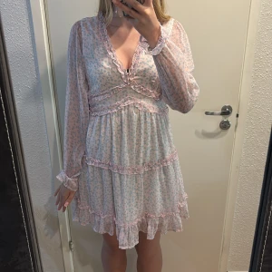 Blommig volangklänning från VERO MODA - Supersöt kort blommig klänning från VERO MODA i ljusa pastellfärger. Klänningen har v-ringning, långa ärmar och flera volangdetaljer med rosa kant. Tunt, luftigt material och småblommigt mönster i blått och rosa. Perfekt för sommarens alla tillfällen!