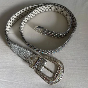 BB belt rhinestone - Snyggt bälte i silver med massor av glittrande strass och nitar längs hela bandet. Spännet är dekorerat med stenar och bältet har en cool, glittrig yta som verkligen sticker ut. Perfekt accessoar för att lyfta vilken outfit som helst.