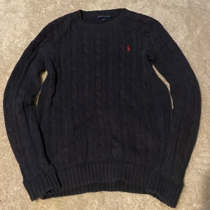 Ralph lauren tröja -  Kabelstickad mörkblå tröja från Ralph Lauren med klassisk rund hals och röd broderad logga på bröstet. Tröjan har långa ärmar och ribbade muddar för en snygg passform. Perfekt för dig som gillar stilren och tidlös design. Tröjan är en M men sitter som en XS