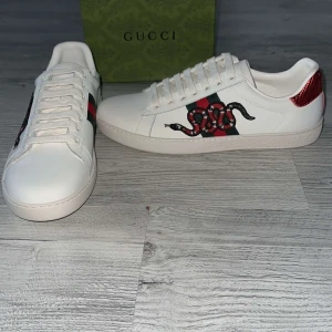 Gucci Ace sneakers med orm  - Gucci Ace sneakers i vitt skinn med ikonisk röd och grön rand samt broderad orm på sidan. Baksidan har metallic-detaljer i rött och grönt med Gucci-logga. Klassisk rund tå, snörning och platt sula. Perfekta för dig som vill sticka ut med lyxig streetstyle.