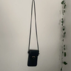 Svart axelväska med hjärt-detalj - Liten svart axelväska i syntet med lång rem och vit kontrastsöm. Framsidan har ett guldfärgat hjärta som stängning och baksidan har en genomskinlig ficka. Perfekt för mobilen och småprylar, stilren och söt design.