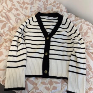 Randig kofta från NA-KD med knappar - Superfin stickad kofta från NA-KD i vitt med svarta ränder och breda ribbade muddar. Koftan har v-ringning, svarta kanter och snygga knappar framtill. Perfekt för lager-på-lager och ger en chill men ändå stilren vibe.