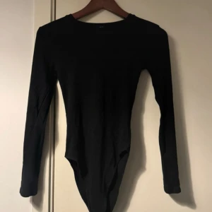 Svart långärmad body från Zara  - En svart, långärmad body från Zara i mjuk bomullsmix. Klassisk rund halsringning och tryckknappar i grenen för enkel på- och avtagning. Perfekt att styla med jeans eller kjol.