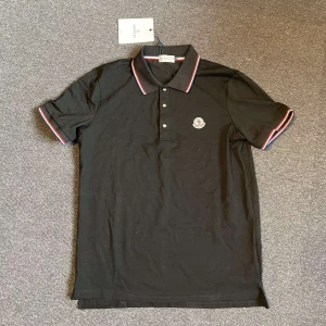 Svart Moncler pikétröja - Svart pikétröja från Moncler med klassisk krage och knäppning framtill. Tröjan har röda, vita och blå ränder på krage och ärmslut samt Moncler-logga broderad på bröstet. Tillverkad i mjuk bomull och har korta ärmar.