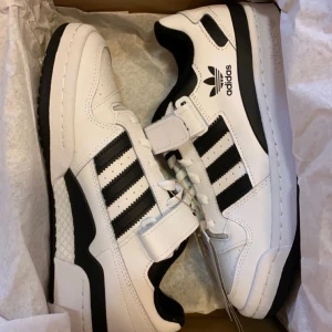 Adidas Forum Low vita och svarta sneakers - Snygga Adidas Forum Low sneakers i vitt och svart med klassiska tre ränder på sidan. Skorna har snörning och kardborreband, rund tå och platt sula. Tillverkade i skinn och syntet, perfekta för en streetwear-look.