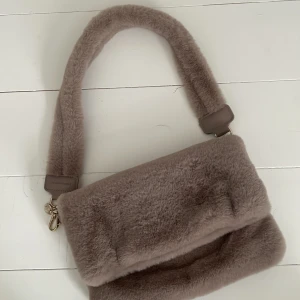 Grå pälsväska från Carin Wester - Supermjuk axelväska i grå fuskpäls från Carin Wester. Väskan har bred axelrem, guldfärgade metalldetaljer och innerfack med dragkedja. Perfekt storlek för det viktigaste och ger en mysig vibe till din outfit. Den har dold magnet stängning och nypris ca 300-400kr. 