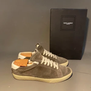 Grå mockasneakers från Saint Laurent - Snygga sneakers från Saint Laurent i grå mocka med vita snören och vit häl. Klassisk låg modell med rund tå och platt sula. Perfekta för dig som gillar stilrena och exklusiva skor med en lyxig känsla.