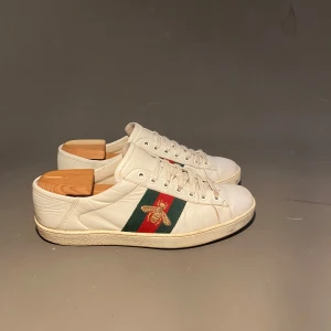 Gucci vita sneakers med bi-detalj - Snygga vita sneakers från Gucci i skinn med klassiska gröna och röda ränder på sidorna. Ena sidan har en broderad guldfärgad bi som ger extra lyxig känsla. Skorna har rund tå, snörning och platt sula. Perfekta för dig som vill sticka ut med stil.