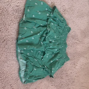 Grön blommig kjol från H&M - Supersöt grön kortkjol från H&M med små vita blommor och bred smock i midjan. Kjolen har volang och är perfekt för en färgglad och lekfull stil. Lätt och luftig, passar perfekt till varma dagar.
