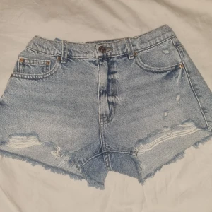 Ljusblå jeansshorts från Gina Tricot - Snygga ljusblå jeansshorts från Gina Tricot med hög/mellanhög midja och råa, fransiga kanter. Shortsen har slitna detaljer framtill, klassiska fickor fram och bak och knappstängning. Perfekta för sommaren. Endast använda 1 gång och i mycket bra kvalité. (Är snäppet mörkare än på bilden då den är tagen med blixt, men ingen större skillnad.) Köp direkt via Plick! Snygga och kan stylas på flera olika sätt inför sommaren!