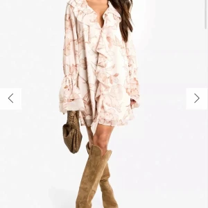 Blommig kortklänning med volang - Supersöt kortklänning i ljusa toner med blommigt mönster och volangdetaljer längs v-ringningen. Klänningen har långa ärmar med volang vid ärmslut och en luftig passform. Perfekt för vår och sommar! Nyskick (prislapp finns kvar på klänningen) 