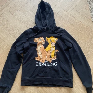 Svart Lion King hoodie XS FB Sister - Svart hoodie från FB Sister i samarbete med Disney Lion King. Framsidan har ett stort tryck med Simba och Nala samt texten 'The Lion King'. Tröjan har huva med snörning och långa ärmar. Perfekt för dig som älskar Disney och vill ha en chill look.