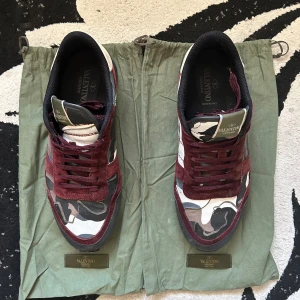 Valentino Rockrunners - Snygga Valentino Rockrunner sneakers med vinröda mockadetaljer och coolt camouflage-mönster i svart, grått, beige och vitt. Skorna har snörning, platt sula och ikoniska nitar i rött på hälen. Perfekta för dig som vill sticka ut med stil.