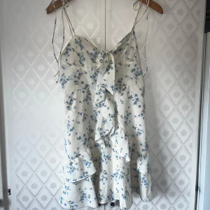 Vit blommig kortklänning med volanger - Supersöt vit kortklänning med blått blommönster och tunna axelband. Klänningen har volanger nertill, omlottvolang framtill och en rosett vid bysten. Perfekt för sommaren. I toppen skick men har små sminkfläckar på banden (se bilder). 