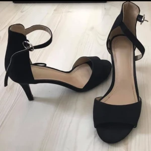 Svarta pumps med öppen tå och klack - Stilrena svarta pumps med öppen tå och smal klack. Skorna har en elegant ankelrem med spänne och är klädda i mjukt material som ger en lyxig känsla. 