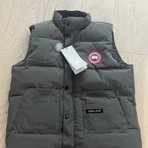 Mörkgrå dunväst från Canada Goose - Säljer en mörkgrå dunväst från Canada Goose med klassisk logga på bröstet och hög krage. Västen har två stora fickor framtill, knappar och dragkedja. Perfekt för lager-på-lager och riktigt snygg till streetwear eller outdoor-stil.