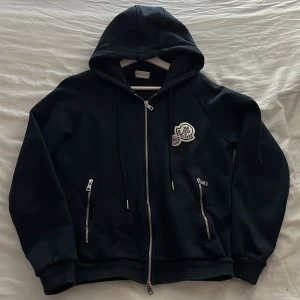 Blå hoodie från Moncler med dragkedja - Blå hoodie från Moncler med huva, tvåvägsdragkedja och två fickor med dragkedjor. Tröjan har Moncler-logga på bröstet och är tillverkad i mjukt material. Perfekt för dig som gillar stilrena och exklusiva plagg.