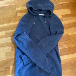 Blå hoodie från Ahlquist Clothing XS - Mysig blå hoodie från Ahlquist Clothing i storlek XS. Tröjan har huva med snörning och långa ärmar. Materialet känns stickat och är perfekt för kyliga dagar. Enkel och stilren design som funkar till det mesta.