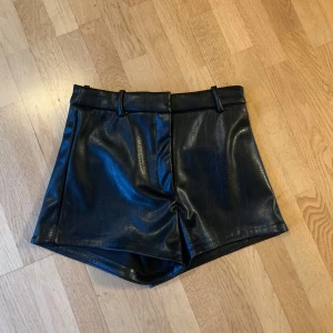 Svarta veganska skinnshorts H&M - Snygga svarta shorts i veganskt skinn från H&M. De har hög midja, bälteshällor och två fuskfickor bak. Perfekt för dig som vill ha en edgy look med stilren känsla. Passformen är normal och materialet är syntetiskt skinn.