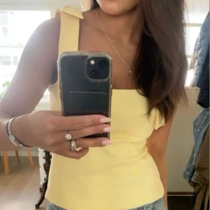 Gul  topp med rosetter - Snygg gul topp med stora rosetter på axlarna. Perfekt för sommardagar och ger en fräsch look. Toppen har en enkel och stilren design som passar till många olika outfits. Nytt skick 