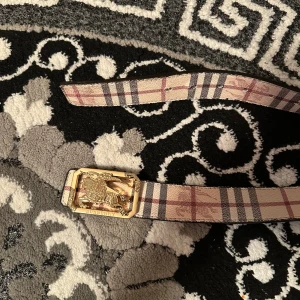 Burberry rutigt bälte med guldspänne - Säljer ett klassiskt Burberry bälte med det ikoniska rutiga mönstret i beige, svart och rött. Bältet har ett snyggt guldigt spänne med Burberry-logga och hästmotiv. Perfekt accessoar för att lyfta din outfit med en lyxig touch.