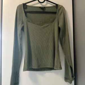 Olivgrön ribbad topp från Monki - Olivgrön långärmad topp från Monki i ribbad bomull med spetsdetaljer vid halsringning och ärmslut. Snygg v-ringning och liten slits vid ärmslutet ger en extra touch. Perfekt att styla med jeans eller kjol för en chill look.