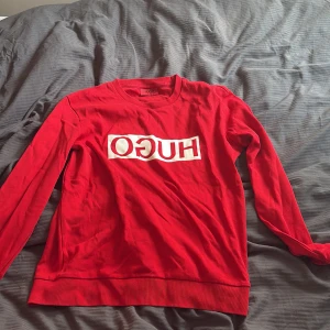 Röd långärmad tröja från Hugo Boss - Röd långärmad tröja från Hugo Boss med stort vitt HUGO-tryck på bröstet. Tröjan har rund halsringning och är gjord i mjuk bomull. Perfekt för dig som gillar en clean och stilren look med en touch av street.