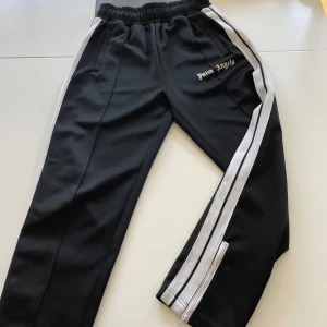 Palm Angels svarta mjukisbyxor med ränder - Svarta mjukisbyxor från Palm Angels med vita sidoränder och logga på vänster höft. Byxorna har elastisk midja, raka ben och dragkedja vid benslut. Snygg sportig vibe och passar perfekt till streetwear-stilen.