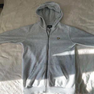 Grå hoodie från Lyle & Scott - Snygg grå hoodie från Lyle & Scott med dragkedja och klassisk gul logga på bröstet. Tröjan har huva, två fickor fram och långa ärmar. Perfekt för chill dagar eller när du vill ha en enkel och stilren look.