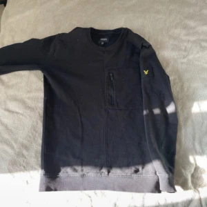  sweatshirt från Lyle & Scott - Svart sweatshirt från Lyle & Scott med rund hals och en bröstficka med dragkedja. Klassisk gul logga på vänster ärm. Tröjan har långa ärmar och är tillverkad i mjuk bomull, perfekt för en chill och stilren look.
