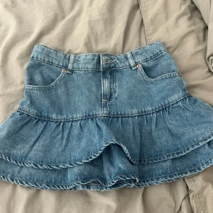 Volangkjol i jeans från Zara - Supersöt blå jeanskjol från Zara med volang och klassiska fickor fram. Kjolen har en kort modell och är tillverkad i denim med snygga sömdetaljer.