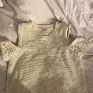 Stickad ärmlös topp med spetsdetaljer - Supersöt stickad topp i ljusbeige med ärmlös design och volangliknande spetsdetaljer vid axlarna. Perfekt för dig som gillar romantisk och bohemisk stil. Rund halsringning och mjukt material som känns skönt mot huden.