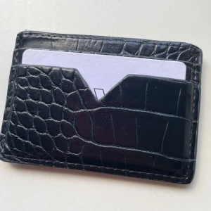 Svart korthållare i konstskinn från Carin Wester - Slimmad svart korthållare från Carin Wester i präglat konstskinn med croco-mönster. Perfekt för dig som vill ha ett stilrent och smidigt sätt att bära dina kort. Snygg och enkel design med plats för flera kort.