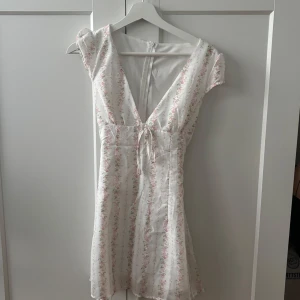 Vit blommig kortklänning med knyt - Supersöt vit kortklänning med små rosa blommor och gröna blad, v-ringning och knyt framtill. Klänningen har dragkedja i ryggen och korta ärmar. Perfekt för sommarens alla tillfällen när du vill känna dig extra fin.