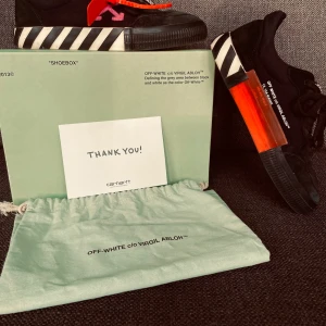 Off-White x Virgil Abloh svarta sneakers - Svarta sneakers från Off-White c/o Virgil Abloh med ikoniska vita ränder på sulan, orange tag och textdetaljer på sidan. Skorna har snörning och en chunky platt sula. Materialmix av mocka och syntet. Perfekt för dig som gillar streetwear och unika detaljer.