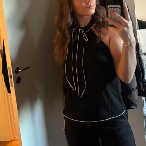 Svart halterneck linne med knytband - Säljer ett svart halterneck linne med vita kanter och snyggt knytband i halsen. Toppen är ärmlös och har en lite loose passform. Använd en gång bara, storlek 36!