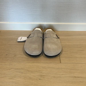 Beige mockasandaler från Birkenstock - Snygga sandaler från Birkenstock i beige mocka med justerbart spänne i metall. Skorna har en platt sula i svart och en klassisk rund tå. Perfekta för dig som gillar stilren och bekväm design. Det står 42 i tofflan, men det är ett designfel! Den riktiga storleken är 39.