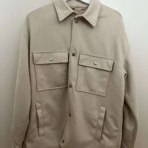 Beige overshirt med stora fickor - Stilren beige overshirt med två stora bröstfickor med lock och knappar. Jackan har klassisk krage, raka ärmar och knäppning framtill. Perfekt för lager-på-lager och enkel att matcha med olika outfits.