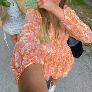 INTRESSEKOLL! Mönstrad kortklänning i orange och rosa - INTRESSEKOLL på kortklänning med färgglatt paisleymönster i orange, rosa och beige toner. Klänningen har v-ringning, långa ärmar med resår och en flowy passform. Perfekt för varma dagar och ger en riktigt somrig vibe!