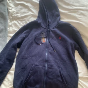 Mörkblå hoodie från Polo Ralph Lauren - Mörkblå hoodie från Polo Ralph Lauren med röd broderad logga på bröstet. Tröjan har huva med snörning, dragkedja framtill och känguruficka. Tillverkad i mjuk bomull, perfekt för chill dagar. Klassisk och stilren design.