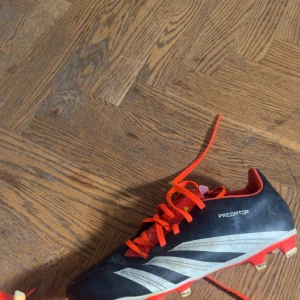 Adidas Predator fotbollsskor svart/röd - Säljer ett par Adidas Predator fotbollsskor i svart med röda och vita detaljer. Skorna har röda snören, klassiska tre ränder på sidan och dobbar för gräsplan. Insidan och sulan är också röda. Perfekta för dig som vill sticka ut på planen.