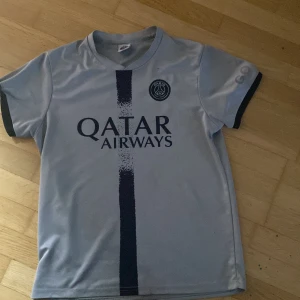 PSG Neymar Jr 10 fotbollströja grå - Snygg PSG fotbollströja i ljusgrått med mörkblå detaljer och tryck. Framsidan har Qatar Airways-logga och PSG-emblem, baksidan Neymar Jr och nummer 10. Tillverkad i lätt och ventilerande material, perfekt för fotbollsträning eller att bära till vardags.