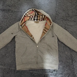 Burberry Zip Up - Grå hoodiejacka från Burberry med klassiskt rutigt foder i huvan. Jackan har dragkedja framtill, kängurufickor och mjukt vitt fleecefoder på insidan. Perfekt för dig som vill ha en stilren look med en touch av lyx och ikoniskt mönster. Storlek M och sitter lite litet som burberry ska göra. Hör av er för eventuella frågor😁. TRÖJAN ÄR ÄKTA, digitalt kvitto följer med påse och tags!!!!