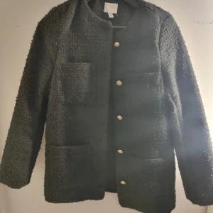 Svart cardigan jacka från H&M XS - Stilren svart cardigan jacka från H&M i storlek XS. Jackan har grov textur, raka axlar, fyra stora fickor framtill och snygga guldiga knappar. Perfekt för dig som gillar klassisk men trendig look. Materialet känns mjukt och har en exklusiv känsla.