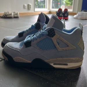 Nike Air Jordan 4 University Blue - Nike Air Jordan 4 University Blue med ljusblå mocka, meshpaneler och detaljer i svart och grått med stänk. Ikonisk Jumpman-logga på hälen och tungan, samt klassisk snörning. Snygg och sportig siluett som sticker ut.